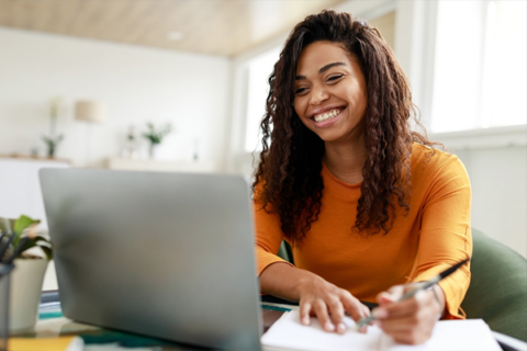 Smiling woman taking notes while working on a laptop.