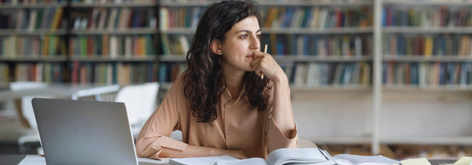 Female student sitting at a desk with a laptop and looking pensively off in the distance.