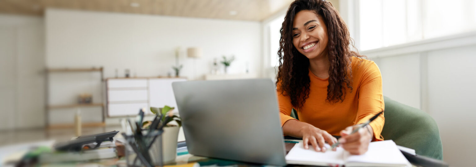 Smiling woman taking notes while working on a laptop.