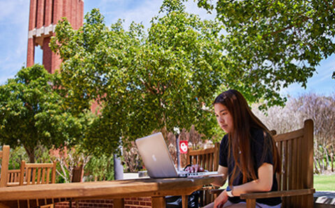 Female OU student sitting at an outside table under blue sky while working on her computer