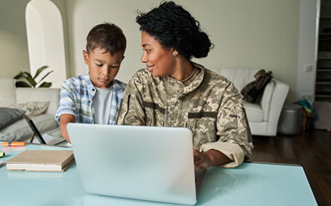 Military student at her home desk with laptop and her young son is standing next to her