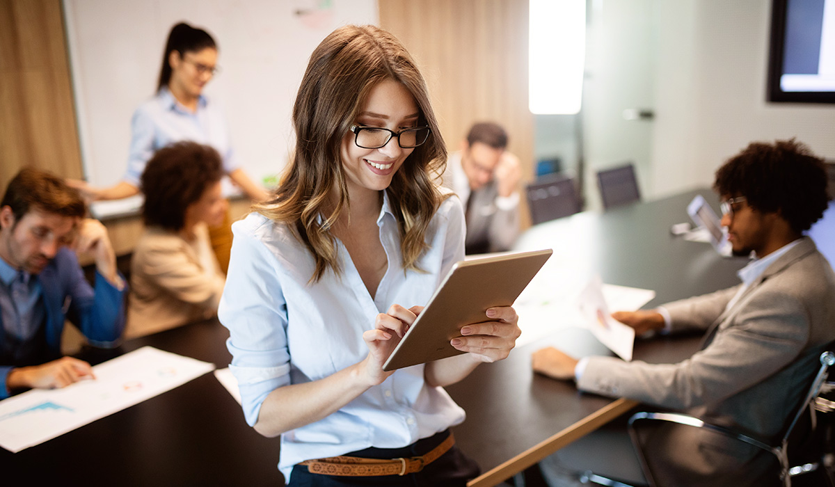 young professional taking notes on a tablet while co-workers are behind her at a conference table