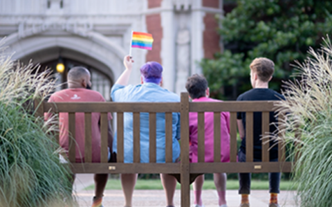 Group of 4 students sitting on a bench in front of Bizzell Library. One student is holding up a small gay pride flag