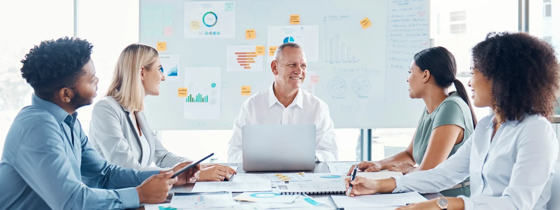 Five data professional sitting at a meeting table with several data documented in the table and on the white board behind them