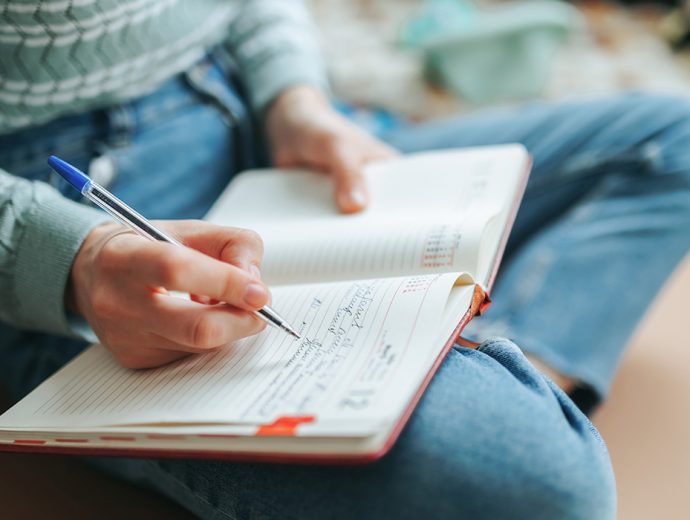 close up of a student sitting on the floor with crossed legs writing in an open notebook that is one her lap